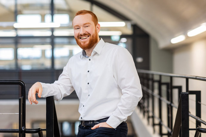 Tom Betz mit rotem Bart und weißem Hemd lächelt, lehnt entspannt an einem schwarzen Geländer.