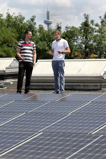 Zwei Personen stehen auf einem Dach mit Solarpaneelen, im Hintergrund sind Bäume und ein Turm zu sehen.