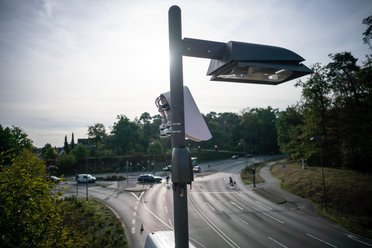 Eine Straßenlaterne mit Kamera und Antenne steht an einer Kreuzung bei Tageslicht.
