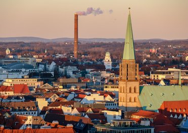 Eine Stadtansicht mit einem Kirchturm im Vordergrund und einem rauchenden Schornstein im Hintergrund bei Sonnenuntergang.