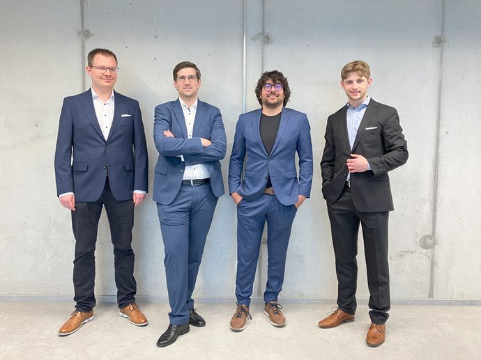 Vier Personen in Anzügen stehen vor einer grauen Wand.