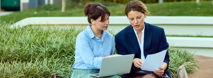 Zwei Personen sitzen draußen, arbeiten gemeinsam mit Laptop und Dokumenten auf einer Bank im Grünen.