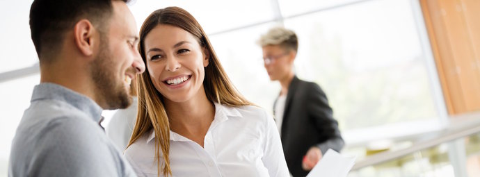 Drei Personen in einem hellen Büro, zwei im Gespräch, eine Person im Hintergrund mit Dokumenten.