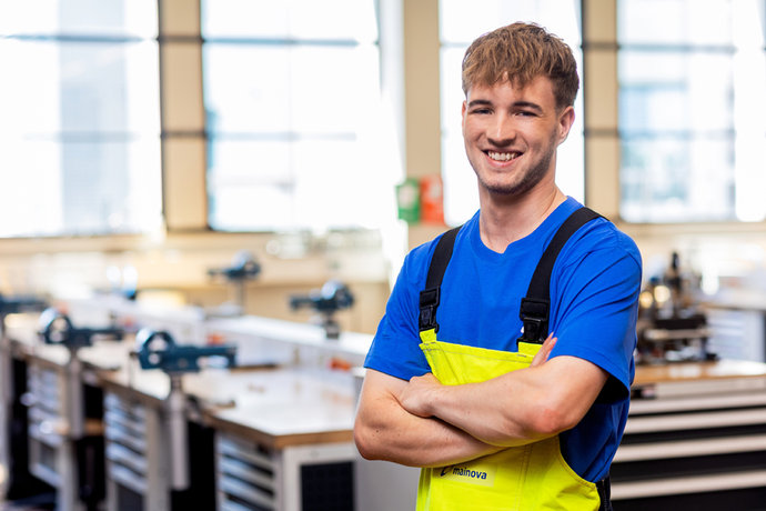 Luca Altoé mit blauem Shirt und gelber Latzhose steht lächelnd in einer Werkstatt.