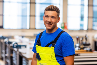 in blauer Kleidung und gelber Latzhose steht lächelnd in einer Werkstatt.