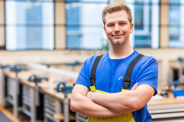 in blauer Kleidung und gelber Latzhose steht lächelnd in einer Werkstatt.