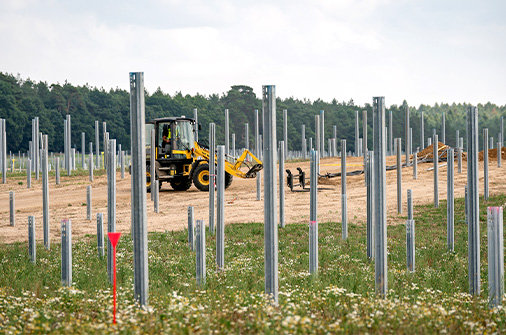 Baustelle eines Solarparks mit vielen gesetzten Metallpfosten und einem Bagger zwischen den Reihen.