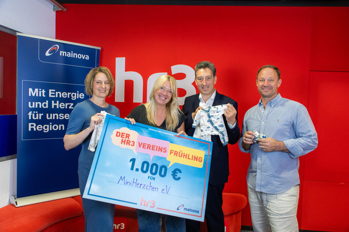 Vier Personen stehen vor hr3‑ und Mainova‑Hintergrund, halten Babykleidung und einen 1.000 Euro‑Spendenscheck.