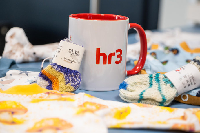Vier Personen stehen vor hr3‑ und Mainova‑Hintergrund, halten Babykleidung und einen 1.000‑Euro‑Spendenscheck.