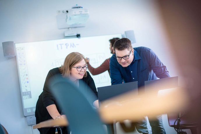 © Mainova/AndreFormer Zwei Auszubildende lehnen sich am Tisch über ein Notebook und betrachten den Monitor