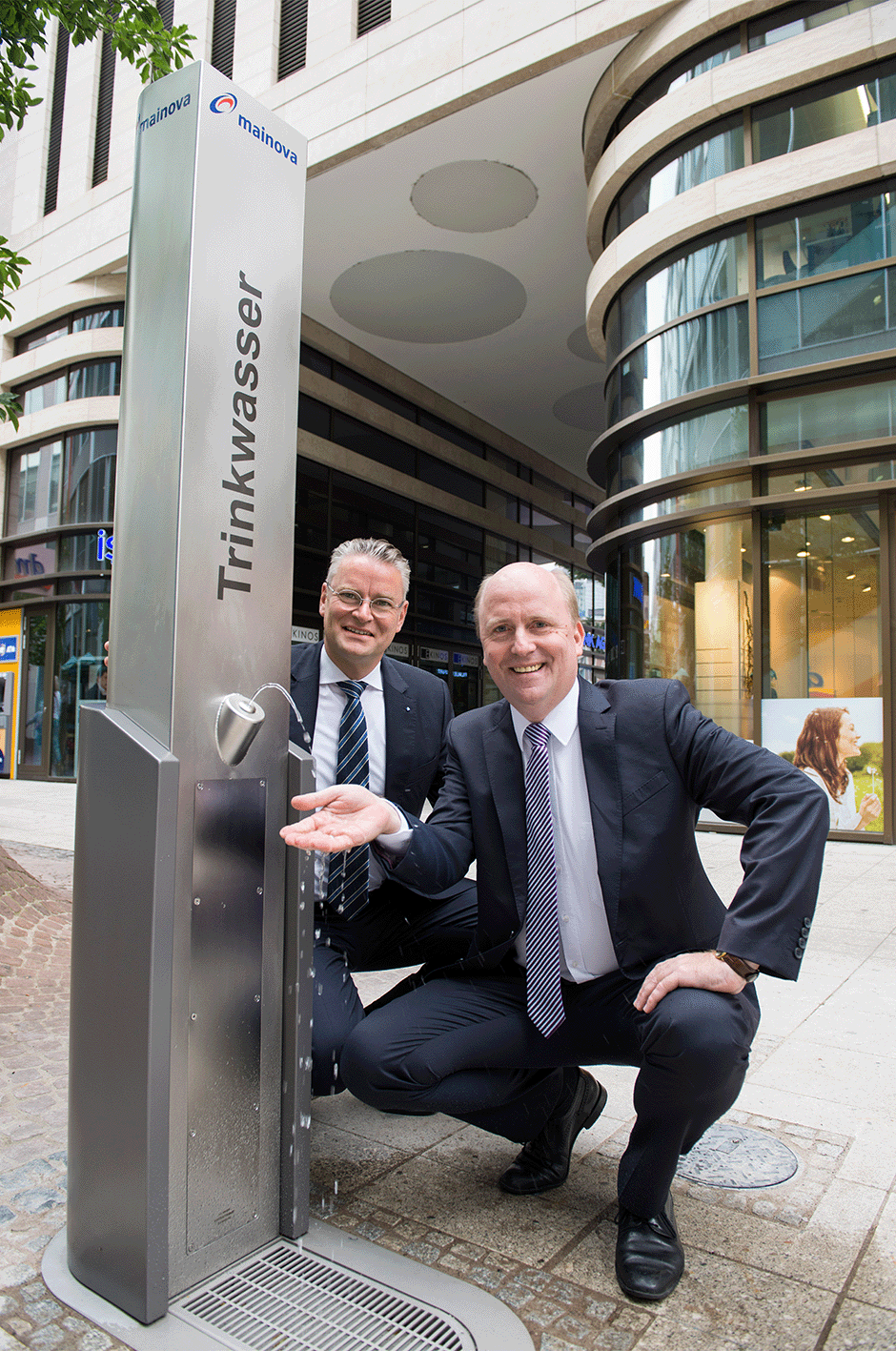 Zwei Männer in Anzügen testen das Wasser aus einem öffentlichen Trinkwasserspender auf einer Straße.