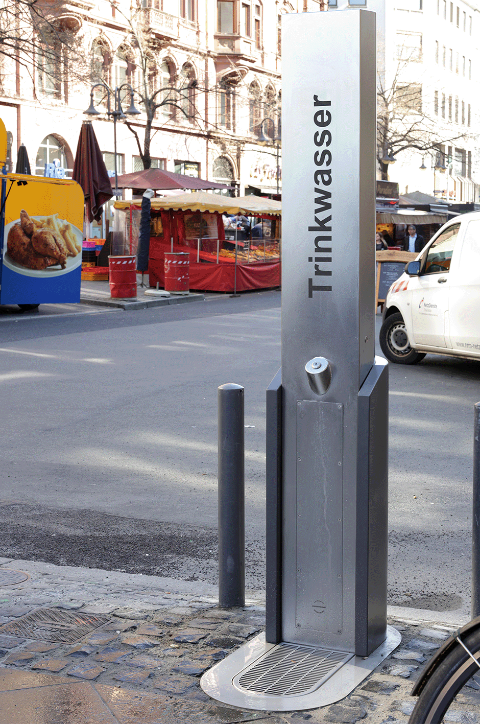 Trinkwasserstation auf einer Straße mit umliegenden Gebäuden und einem Imbissstand im Hintergrund.