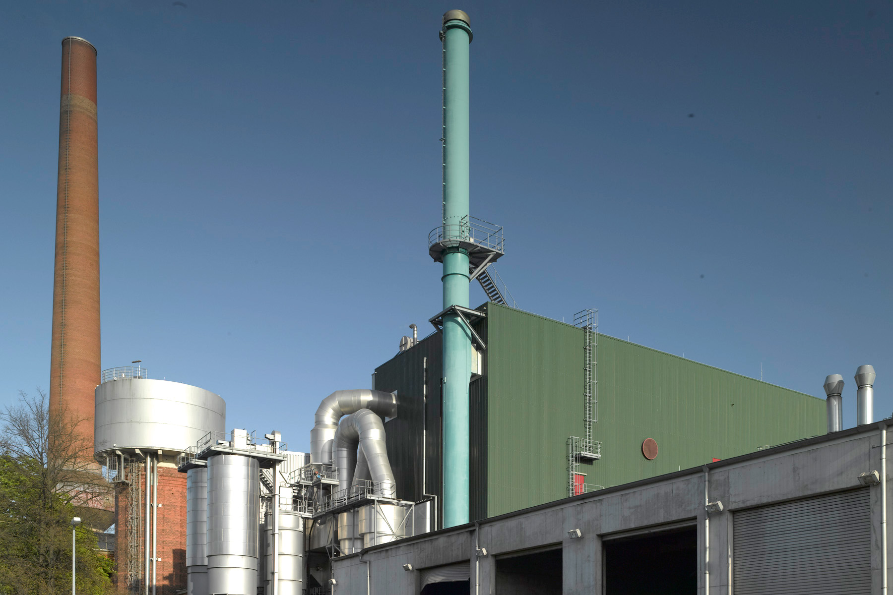 Eine Industrieanlage mit Schornsteinen, Rohren und einem großen grünen Gebäude vor blauem Himmel.