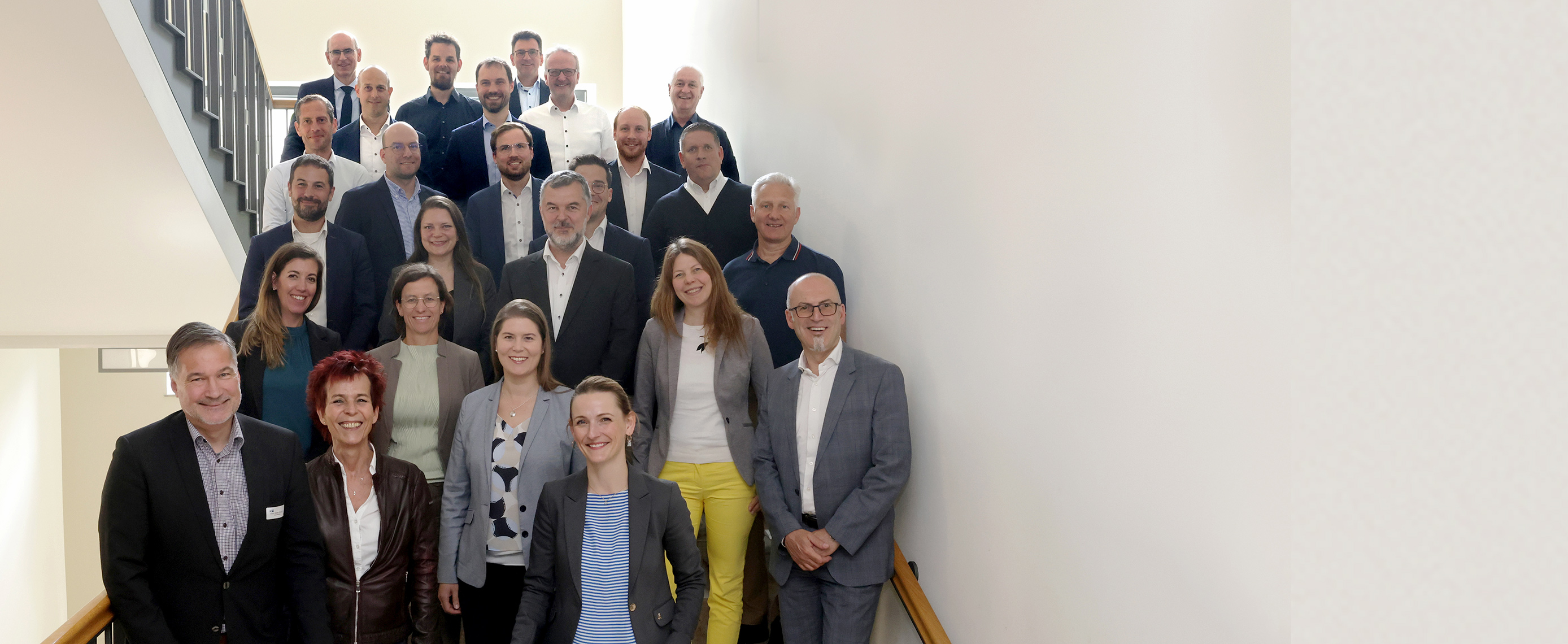 Gruppe von 25 Personen steht lächelnd auf einer hellen Treppe. Sie tragen formelle Kleidung, und der Hintergrund ist schlicht