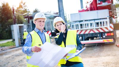 Mann und Frau mit Helm stehen mit Zeichnung am Baufahrzeug. Sie zeigt auf ein Gegenstand außerhalb des Bildes,