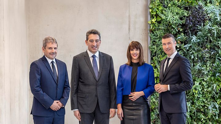 Vier Personen in formeller Kleidung stehen nebeneinander in einem modernen Raum mit einer Pflanzenwand.