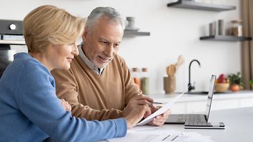 Ein älteres Ehepaar sitzt vor einem Laptop und liest gemeinsam ein schriftliches Dokument, welches der Mann in der Hand hält.