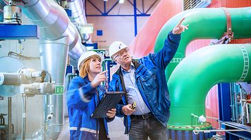 Frau und Mann mit Schutzhelm betrachten Leitungen an der Decke, Und er zeigt auf etwas außerhalb des Bildes