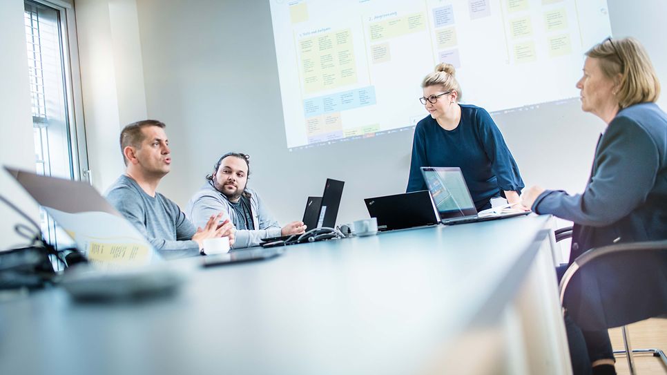 Gruppe sitzt am Konferenztisch und es wird über eine Beamerfolie gesprochen
