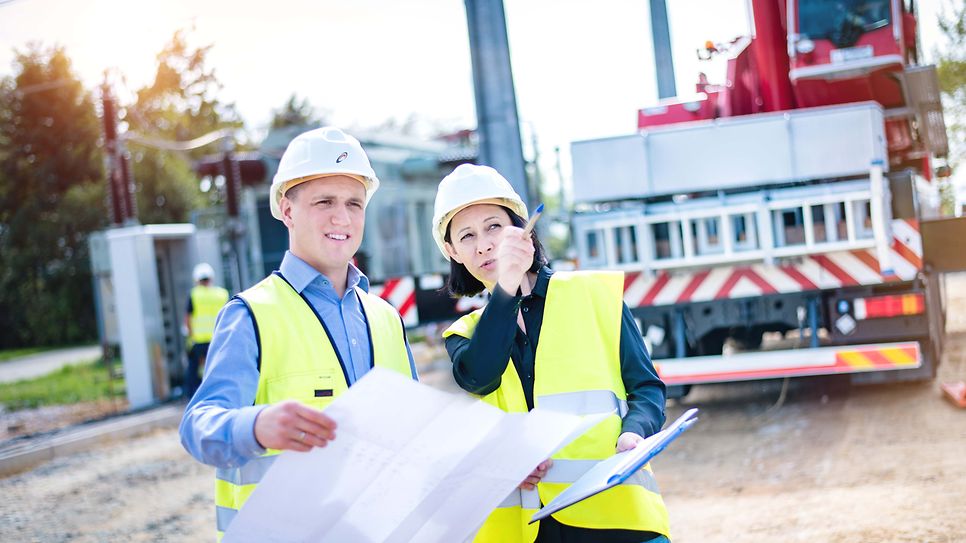 Mann und Frau mit Helm stehen mit Zeichnung am Baufahrzeug. Sie zeigt auf ein Gegenstand außerhalb des Bildes,