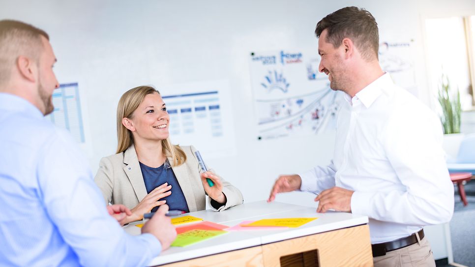 Gruppe steht am Tisch und die Mitarbeiterin erklärt ein Sachverhalt