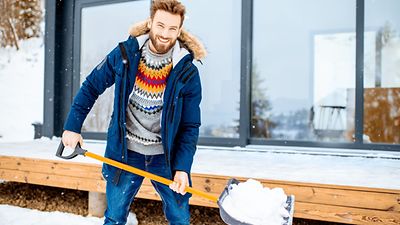 Mann in Winterkleidung schaufelt Schnee und lacht in die Kamera