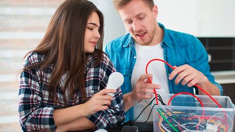 Eine junge Frau und ein junger Mann mit einer Glühbirne und mit Stromkabeln, die er aus einer Kiste nimmt