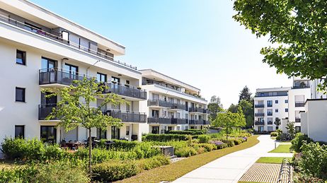 Moderne Wohnanlage mit mehreren weißen Gebäuden, Balkonen und gepflegten Grünflächen bei sonnigem Wetter.