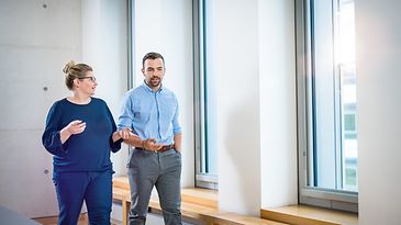 Frau und Mann laufen vor Fensterwand und sprechen miteinander