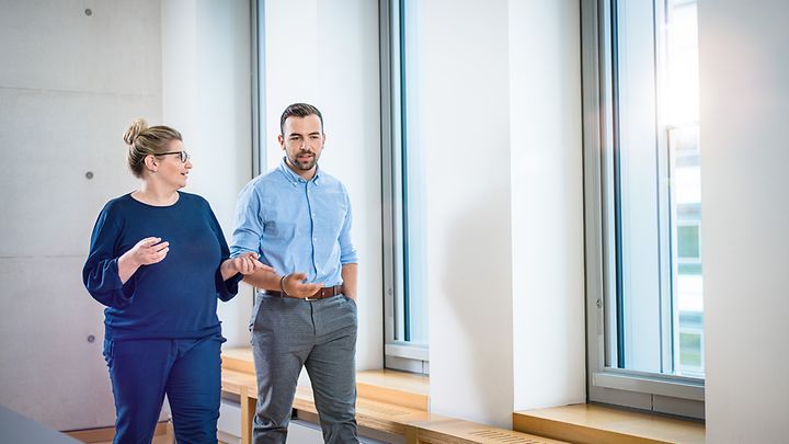 Frau und Mann laufen vor Fensterwand und sprechen miteinander