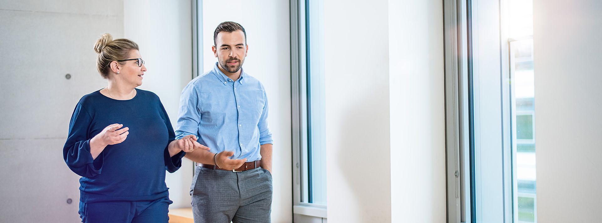 Frau und Mann laufen vor Fensterwand und sprechen miteinander