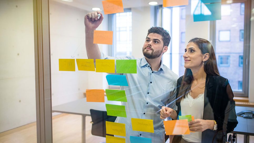 Zwei Studierende hängen Klebezettel an eine Glaswand