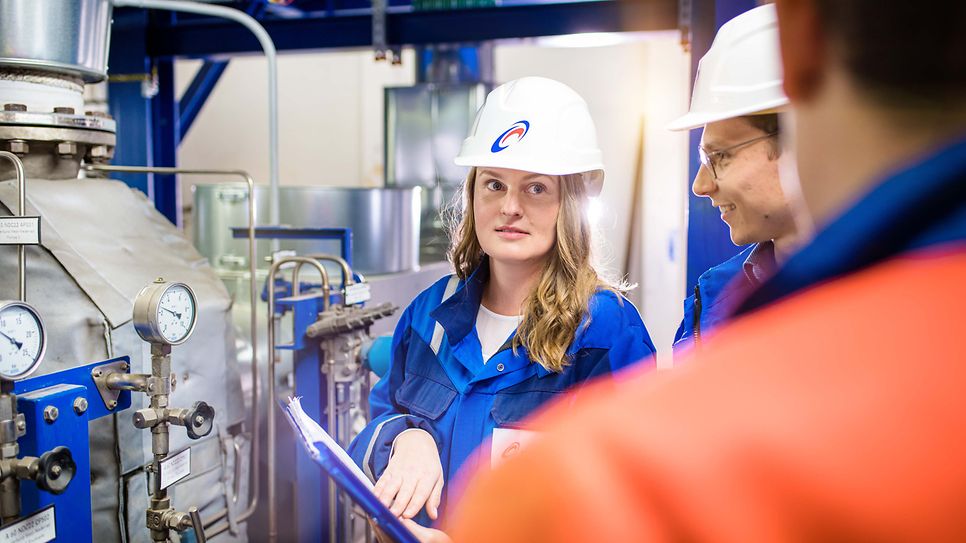 Junge Ingenieurin mit Helm hört ihren Kollegen vor einer Maschine zu