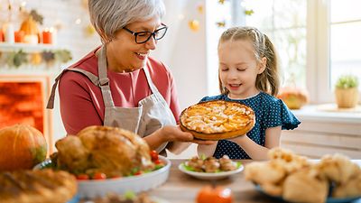 Oma und Enkelin halten einen frisch gebackenen Apfelkuchen, im Vordergrund ein Tisch mit weiteren Speisen