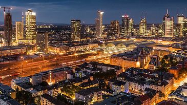 Beleuchtetes Frankfurt in der Nacht: Skyline im Hintergrund, Bahnhof und Zuggleise in der Mitte und Wohnquartier vorne.