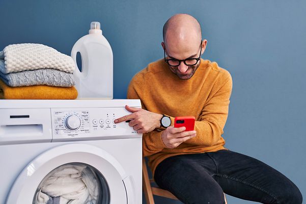 Mann mit Brille schaut auf sein Handy während er seine Waschmaschine bedient.