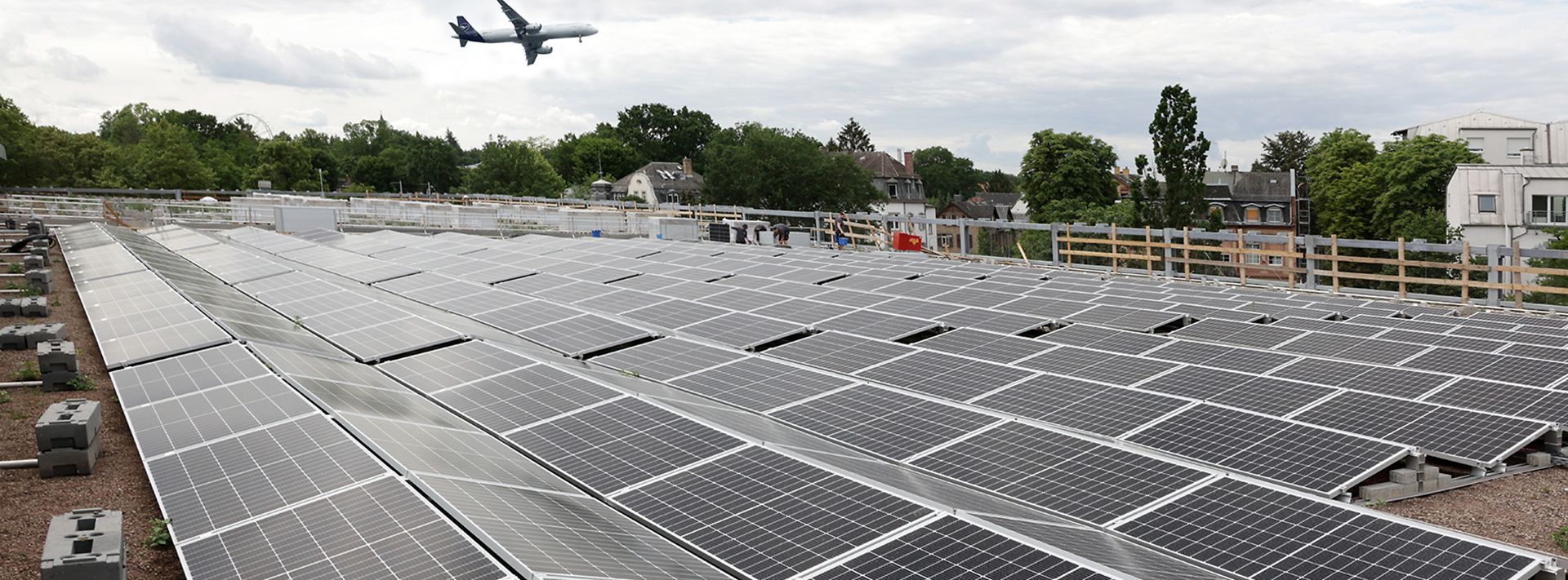 Dach mit vielen Solarpaneelen, im Hintergrund fliegt ein Flugzeug über grüne Landschaft.