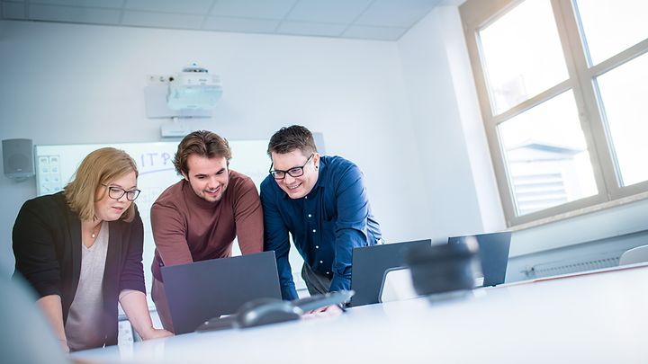 Drei Studierende lehnen sich über Notebook und schauen auf den Monitor