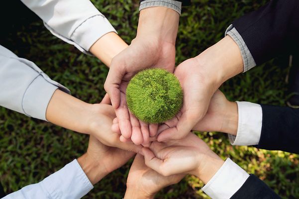 8 Hände ineinander gelegt und von oben fotografiert. Sie halten gemeinschaftlich eine Weltkugel aus Moos