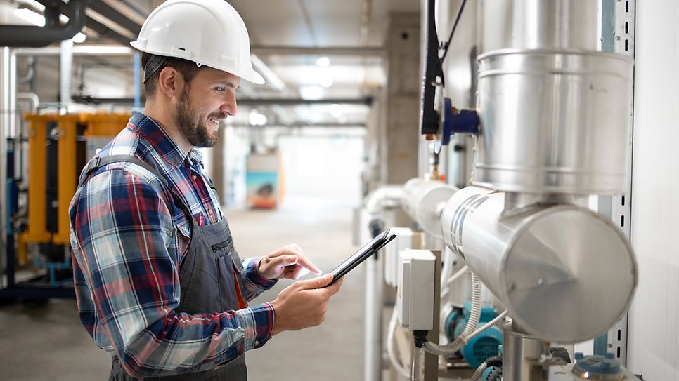 Installateur im Heizungraum steht an den Leitungen und prüft mit dem Tablett die Werte