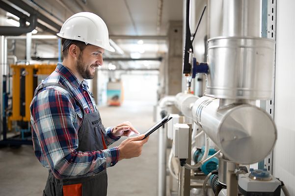 Installateur im Heizungraum steht an den Leitungen und prüft mit dem Tablett die Werte