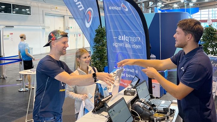 Drei Personen am Messestand lächeln und begrüßen sich, umgeben von Technik.