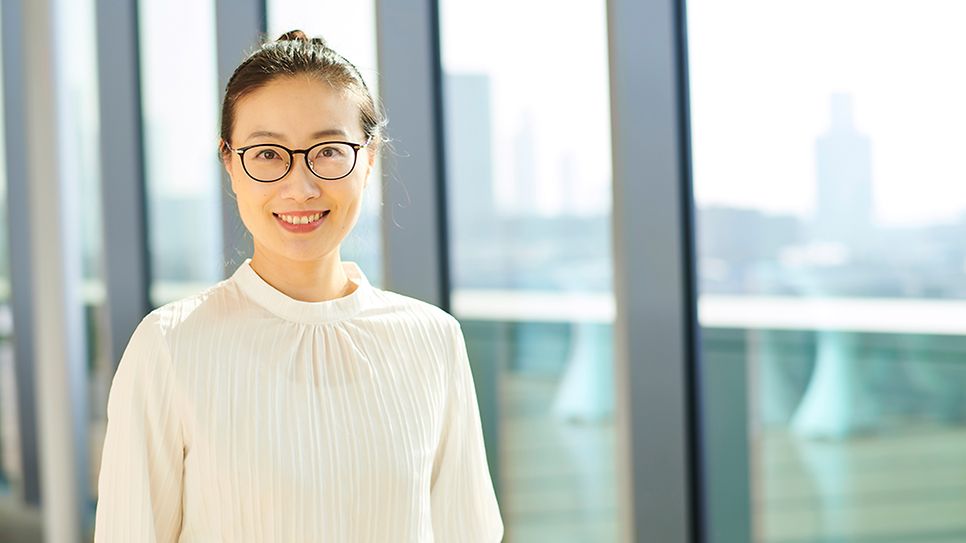 Frau mit Brille lächelt in hellem Raum, weißes Oberteil, Fenster mit Stadtblick dahinter.