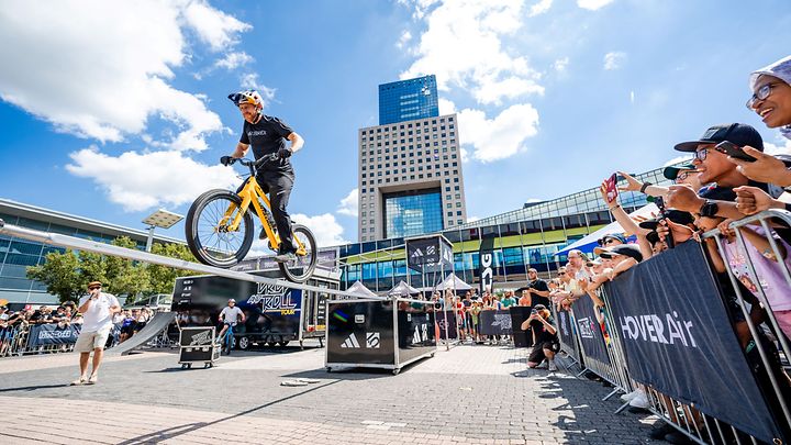 BMX-Fahrer springt über Hindernisse bei Show auf einem Platz, Publikum steht hinter Absperrungen und schaut zu.