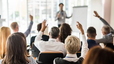 Personengruppe in einem Raum versammelt, stellt Fragen an eine Person die mit einem Flipchart eine Präsentation hält 