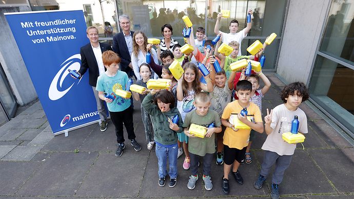 Eine Gruppe von Kindern mit gelben Brotboxen, vier Erwachsene und ein Mainova Aufsteller im Hintergrund