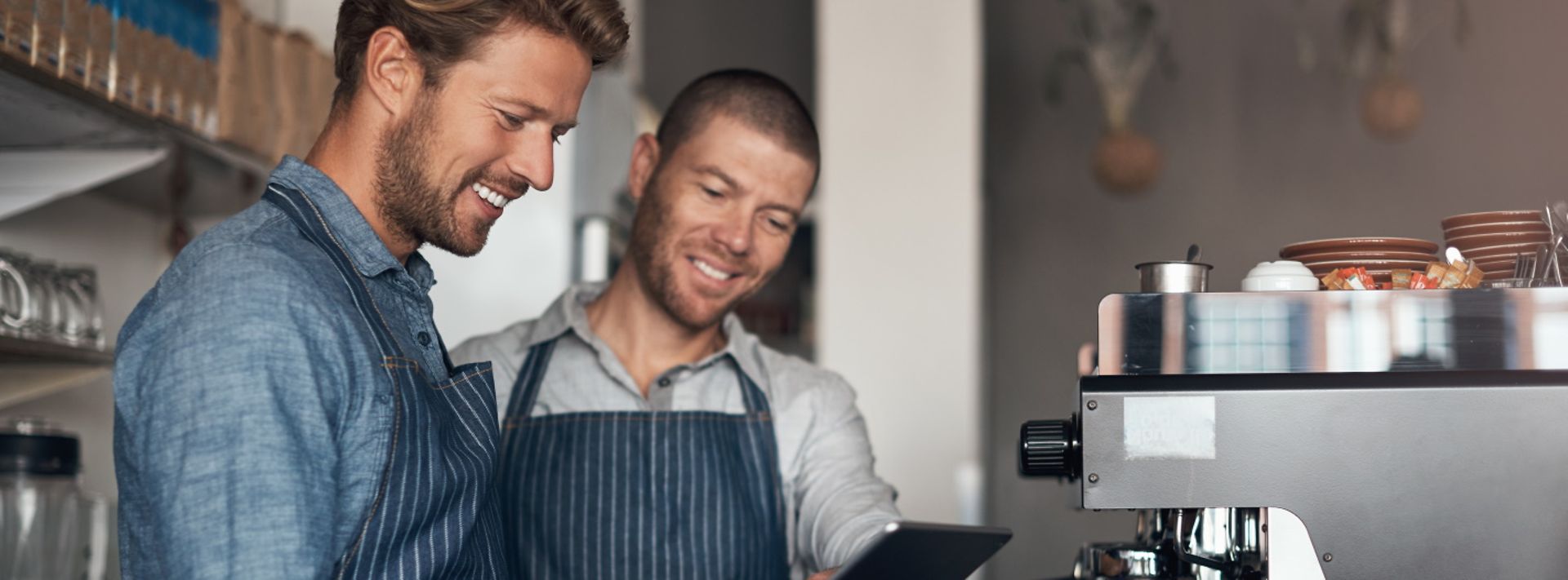 © istockphoto.com/shapecharge Zwei Mitarbeiter in einem Café stehen vor einer Kaffeemaschine und schauen gemeinsam auf ein Tablet.