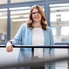 © Mainova/DennisMöbus Eine junge Frau mit Brille und welligem Haar lehnt lächelnd am Geländer in einem modernen Büro mit Glaswänden.