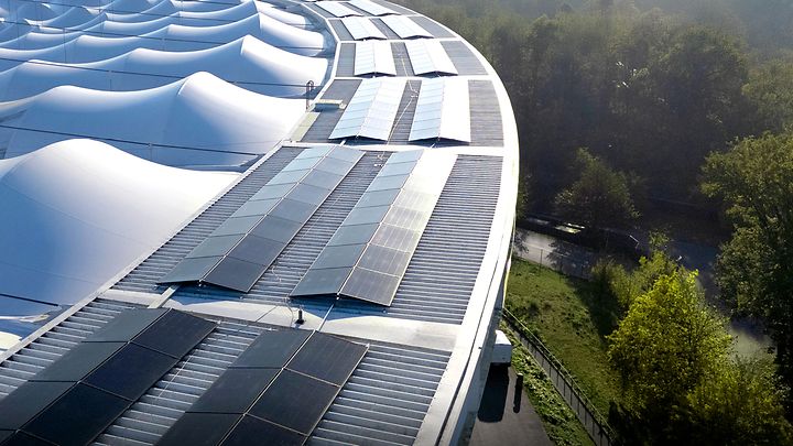 Solarmodule auf einem modernen, gewölbten Dach neben einer grünen Baumlandschaft bei Sonnenlicht aus der Vogelperspektive.