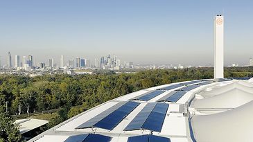 Solarmodule auf einem geschwungenen Dach, dahinter Frankfurts Skyline und viel Grün bei klarem Himmel im Panorama.